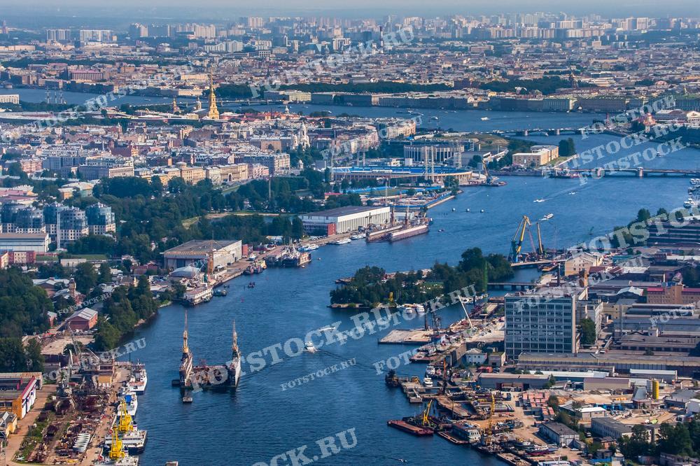 На фотографии изображена панорама Санкт-Петербурга с высоты, с видом на реку Нева, Петропавловскую крепость и центральные районы города. Видны портовые сооружения и множество исторических зданий. Фото может использоваться для туристических буклетов, открыток и рекламных материалов.